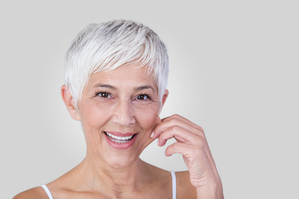 Senior woman pulling cheeks to feel softness and looking at camera. Beauty portrait of happy mature woman smiling with hands on cheek isolated over grey background. Aging process and skin concept. - Foto, Imagen
