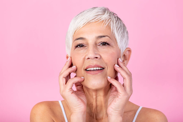 Portrait of beautiful senior woman touching her perfect skin and looking at camera. Closeup face of mature woman with wrinkles massaging face isolated over pink background. Aging process concept. - Photo, Image