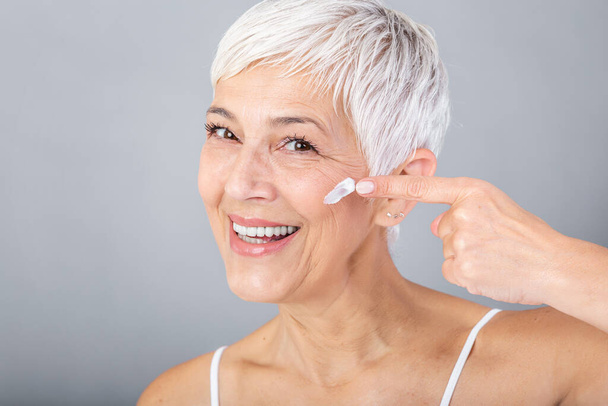 Smiling senior woman applying anti-aging lotion to remove dark circles under eyes. Mature woman using cosmetic cream to hide wrinkles. Lady using day moisturizer to counteract the aging of the skin. - Photo, Image