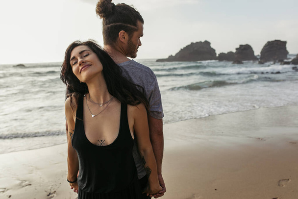 татуированная женщина и бородатый мужчина стоят спиной к спине и держатся за руки возле океана - Фото, изображение