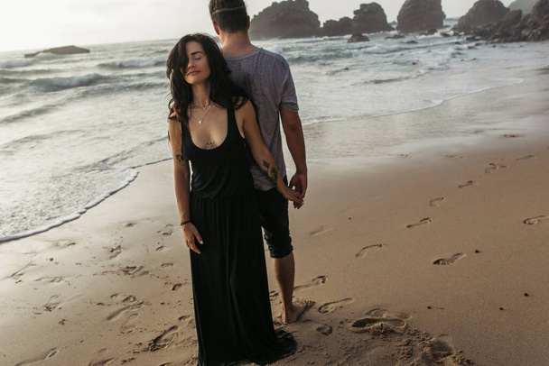 full length of tattooed woman in dress and man standing on beach and holding hands near ocean - Foto, Imagem
