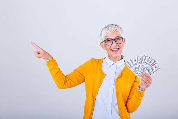 Happy senior woman holding lots of cash, elderly woman with one hundred dollar bills on white background and pointing with her finger to the side copy space. - Foto, imagen