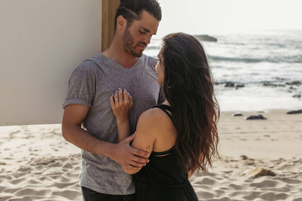 barbudo hombre abrazando tatuado novia con el pelo ondulado en playa cerca del océano - Foto, Imagen