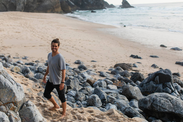 hombre feliz y barbudo en camiseta de pie cerca de piedras en la playa cerca del océano - Foto, imagen