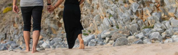 cropped view of couple holding hands and walking on sand near rocks, banner - Фото, изображение