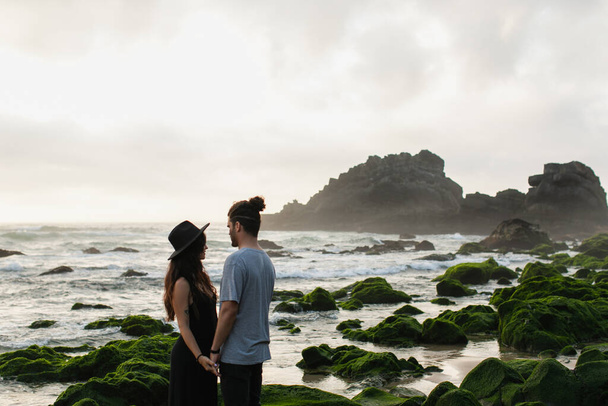 woman in hat and dress holding hands with bearded boyfriend near mossy stones and ocean - Photo, Image