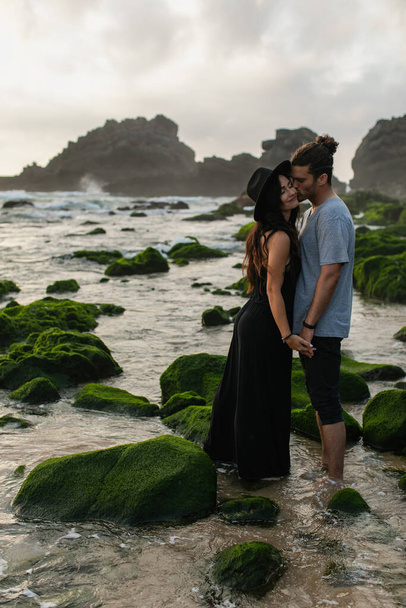 бородатый мужчина целует щеку подружки в шляпе и платье рядом с мхом камни и океан - Фото, изображение