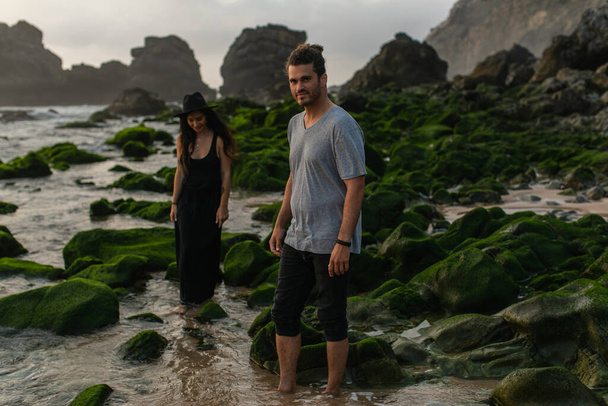 bearded man and tattooed woman in dress and hat walking near mossy stones and  ocean - Foto, Bild