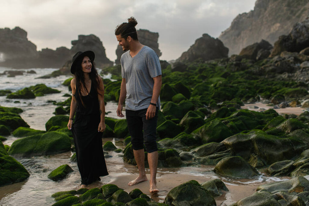 hombre feliz y mujer tatuada en vestido y sombrero caminando cerca de piedras musgosas y el océano - Foto, imagen
