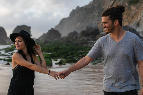 pleased woman in hat and dress holding hands with bearded boyfriend near ocean - Fotó, kép