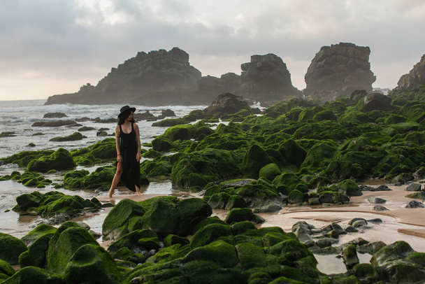 teljes hossza tetovált nő fekete ruha és kalap áll közelében moha kövek óceán - Fotó, kép