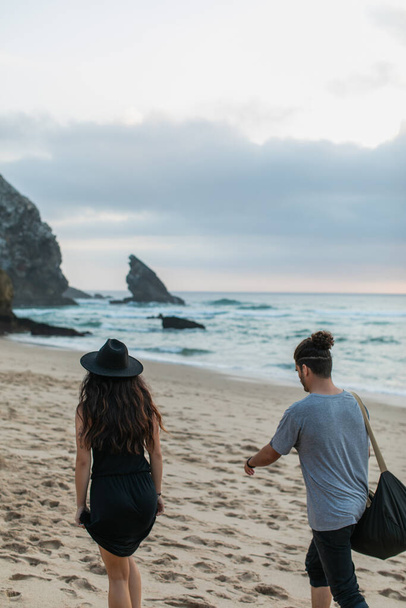 zadní pohled na tetovanou ženu a vousatého muže s taškou kráčející společně na písečné pláži - Fotografie, Obrázek