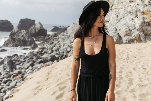 getatoeëerde vrouw in zwarte jurk en hoed staande op zandstrand in portugal  - Foto, afbeelding