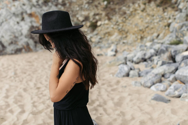 tattooed woman in dress and black hat standing on sandy beach in portugal  - Фото, зображення