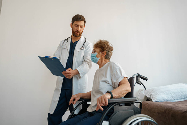 Doctor in mask explaining to a sick patient in wheelchair details of treatment. High quality photo - Фото, изображение