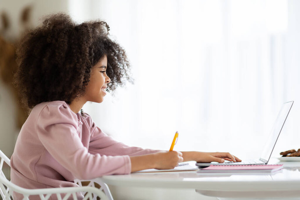 Side view of smiling african american little girl typing on laptop keyboard, schooler doing homework, cute preteen kid working on educational project, copy space. Home schooling, e-education concept - Valokuva, kuva