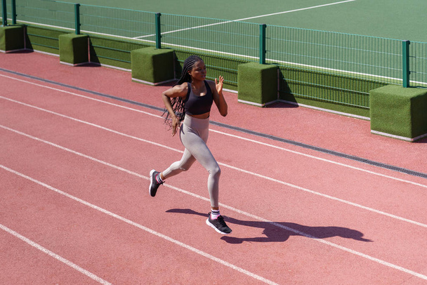 Young black sportswoman enjoys running on red track around stadium to improve technique and increase endurance. African American woman with long braids prepares for upcoming marathon on hot summer day - Fotoğraf, Görsel