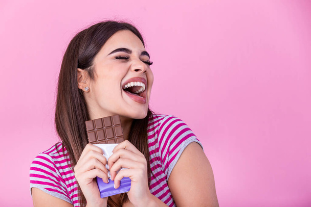 Happy young beautiful lady eating chocolate and smiling. Girl tasting sweet chocolate. Young woman with natural make up having fun and eating chocolate isolated on pink background - Фото, изображение