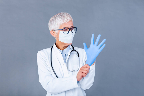 Mature female Doctor putting on protective gloves and medical protective mask, isolated on background. Doctor putting on sterile gloves - Foto, Imagen