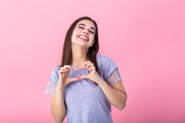 Portrait of a smiling young beautiful woman showing heart gesture with two hands and looking at camera isolated over pink background - Photo, image