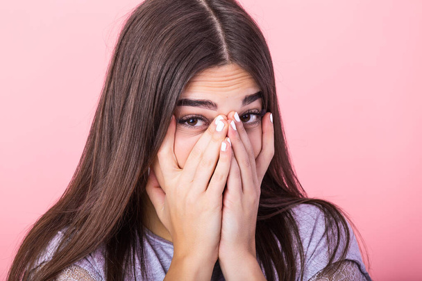Photo of frightened woman with brown hair in basic t-shirt keeping hands over her face and looking at camera isolated over pink background - Valokuva, kuva