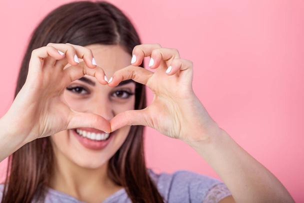 Close-up portrait of cheerful attractive woman showing heart gesture, looking at camera, isolated on pink background - Foto, imagen