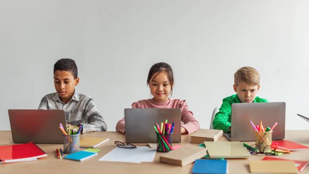 Three Multiracial School Kids Using Laptops Learning Online Sitting At Desk In Modern Classroom At School. Schoolboys And Schoolgirl Having Class Browsing Internet On Computers. Panorama - Φωτογραφία, εικόνα