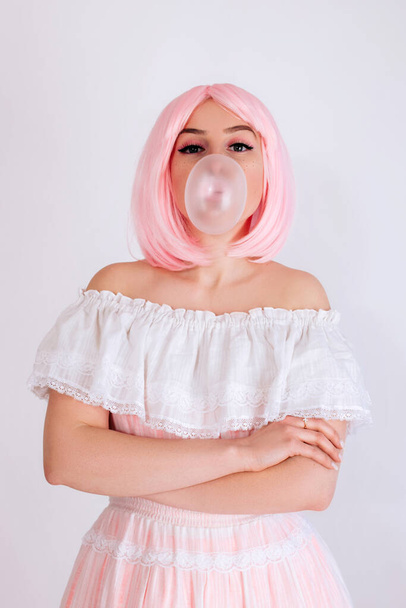 A girl with pink hair, with freckles and in a pink dress inflates a chewing bubble isolated on a white background. A girl with pink makeup blows gum. - Photo, Image
