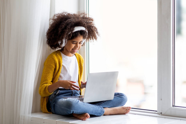Closeup of cheerful black little girl with wireless headset using laptop at home, sitting on window and looking at screen, gesturing and smiling, having video conference with friends, copy space - Foto, afbeelding