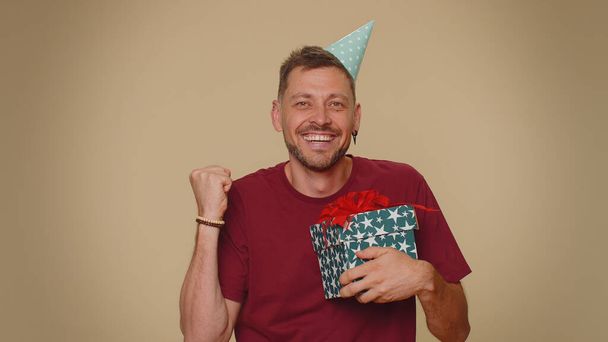 Cheerful bearded handsome young man receiving gift box and smiling joyfully, looking amazed satisfied with nice present, unexpected birthday surprise. Stylish male guy on beige studio background - 写真・画像