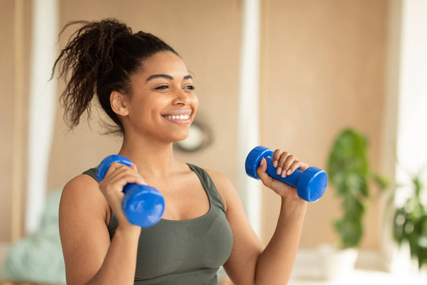 Fitness concept. Portrait of happy african american lady exercising with two dumbbells and smiling, enjoying domestic workout, free copy space. - Foto, afbeelding