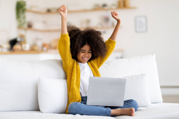 Happy african american child preteen girl sitting on couch at home, using modern laptop, raising hands up and smiling, celebrating success, playing educational games on computer, copy space - 写真・画像
