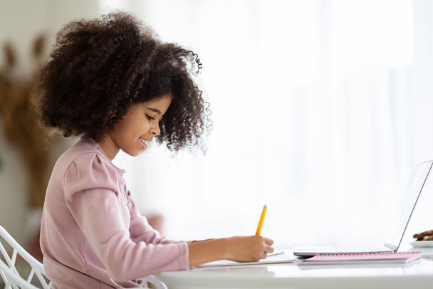 SIde view of adorable little black girl with cute bushy hair doing homework, sitting at desk in front of computer, taking notes, home interior, copy space. Kids education concept - Foto, Imagen