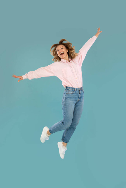 Vertical Shot Of Joyful Woman Posing In Mid Air Having Fun Over Blue Background. Carefree Female Jumping In Studio. Freedom And Happiness Concept. Full Length - Fotografie, Obrázek