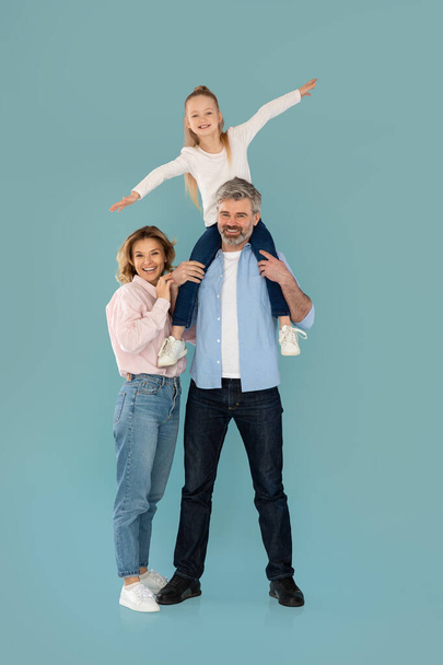 Cheerful Family Posing Together Having Fun Smiling To Camera, Father Holding Kid On Shoulders On Blue Background. Vertical Shot Of Parents And Daughter Standing In Studio - Фото, изображение