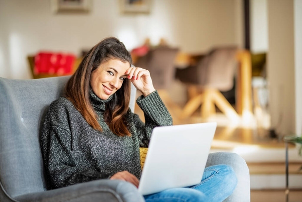 Happy woman using laptop and browsing on the internet while relaxing in the armchar at home. - Valokuva, kuva