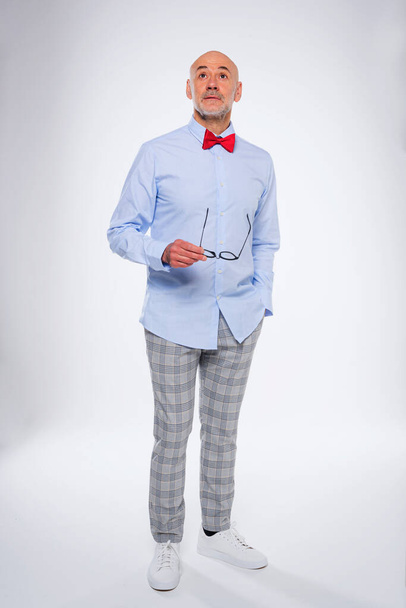 Full length of wondering caucasian mature man. Confident male is wearing shirt and bow tie. He is holding eyeglasses in his hand while standing at isolated background. - Valokuva, kuva