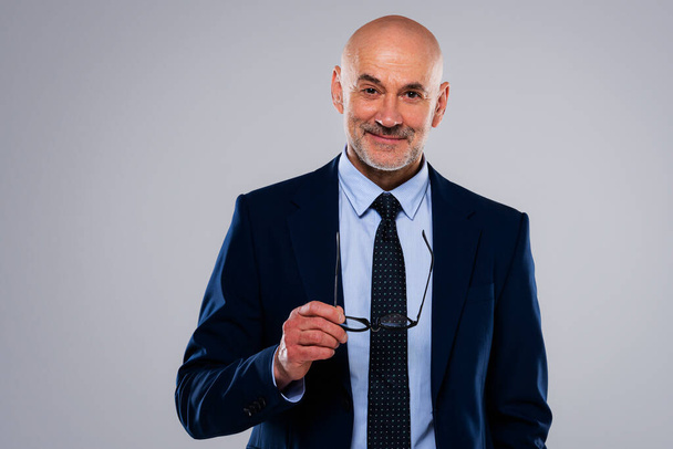 Portrait of smiling caucasian mature businessman. Confident male professional is wearing blazer while holding eyewear in his hand.. He is against gray background. - Foto, immagini