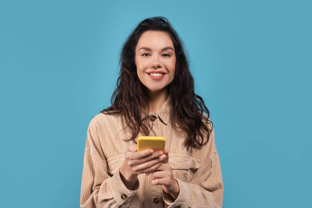 Portrait of happy millennial caucasian brunette woman in casual typing on smartphone, isolated on blue background, studio, empty space. Great ad and offer, social media chat, new app and good message - 写真・画像