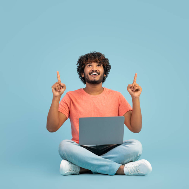 Positive millennial indian man in casual outfit sitting on floor, using modern laptop on blue studio background, typing on computer keyboard, working online, pointing up at copy space - Fotografie, Obrázek