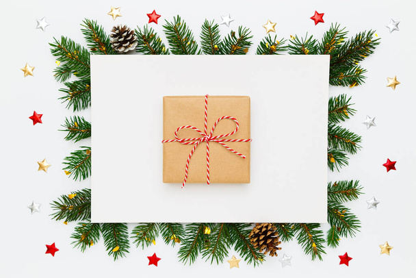 Christmas, New Year holiday layout. Christmas gift in craft paper in frame of natural fir tree branches and pinecones, decorations, stars on white flat lay background - Zdjęcie, obraz