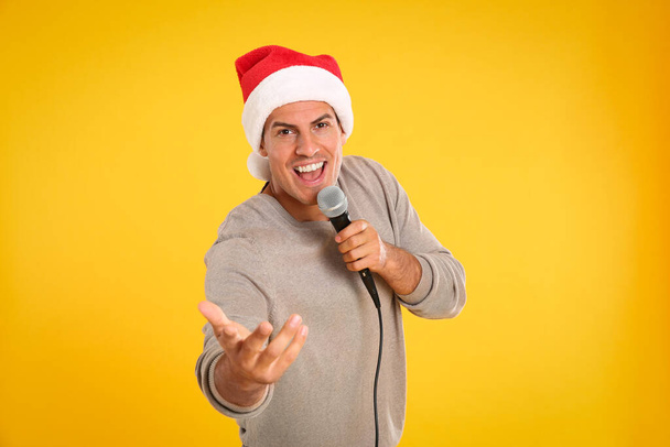 Szczęśliwy człowiek w kapeluszu Świętego Mikołaja śpiewa z mikrofonem na żółtym tle. Muzyka świąteczna - Zdjęcie, obraz