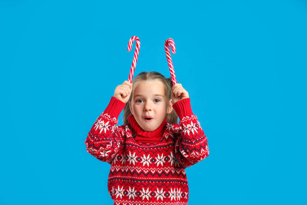 Surprised little girl in knitted Christmas sweater holding candy canes on blue background - Фото, зображення