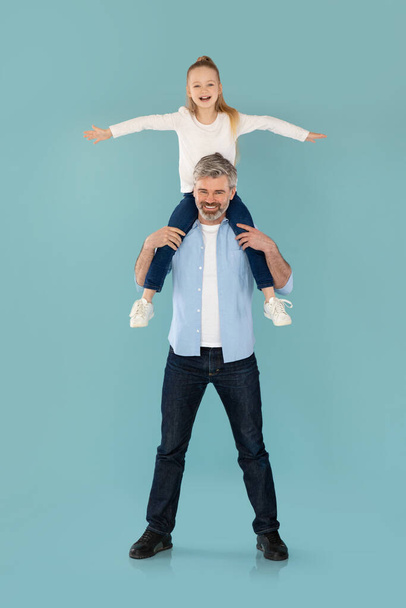 Vertical Shot Of Daddy Holding Little Daughter On Shoulders Posing And Having Fun Over Blue Studio Background, Smiling To Camera. Family And Parenthood Concept. Full Length - Photo, image