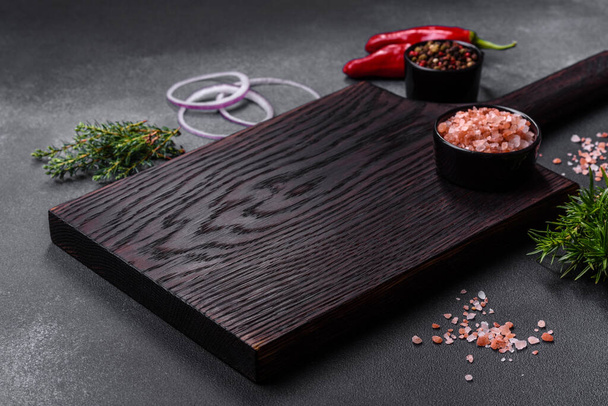 Wooden cutting board, salt, pepper, spices and herbs on a dark concrete background. Cooking delicious home-cooked food - Foto, Imagen