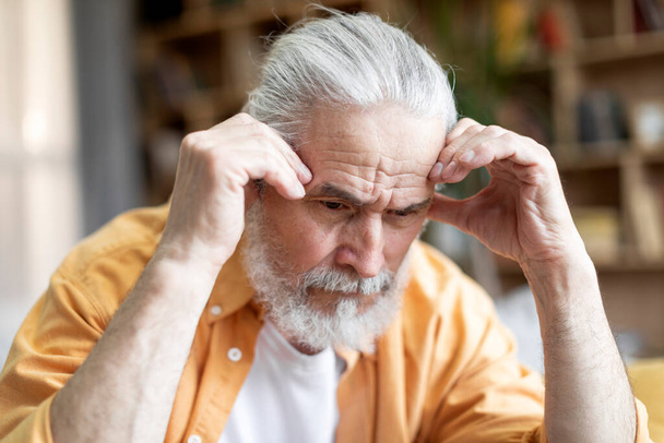 Closeup portrait of sad bearded grey-haired senior man in casual outfit sitting on couch alone at home, touching his head, looking down, feeling broken, having heavy thoughts - Фото, изображение