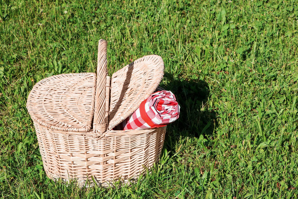 Rolled checkered tablecloth in picnic basket on green grass outdoors, space for text - Photo, image