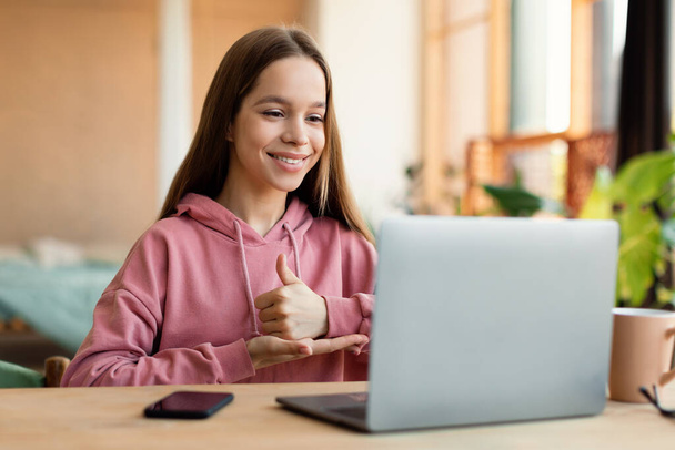 Distant education. Friendly teen girl having online lesson via laptop and showing thumb up on open palm, gesturing in sign language, studying online from home - Photo, Image