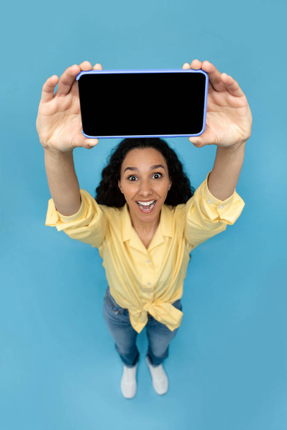 Portrait of cheery young woman holding smartphone with blank screen, presenting gadget with free space for mockup, recommending cool mobile app or website, blue background. Above view - Fotó, kép