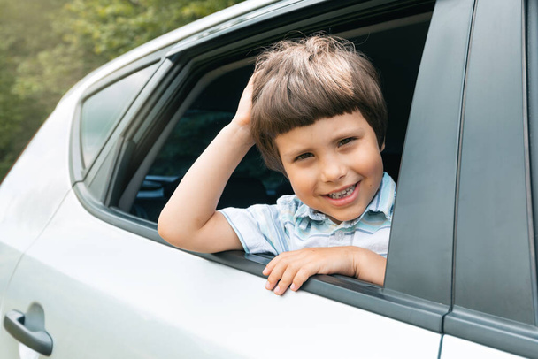 Cheerful caucasian small kid go on vacation by car and looks out window, enjoys trip and weekend, outdoor, close up. Family journey on new car together, safety in auto and life insurance, copy space - Photo, image